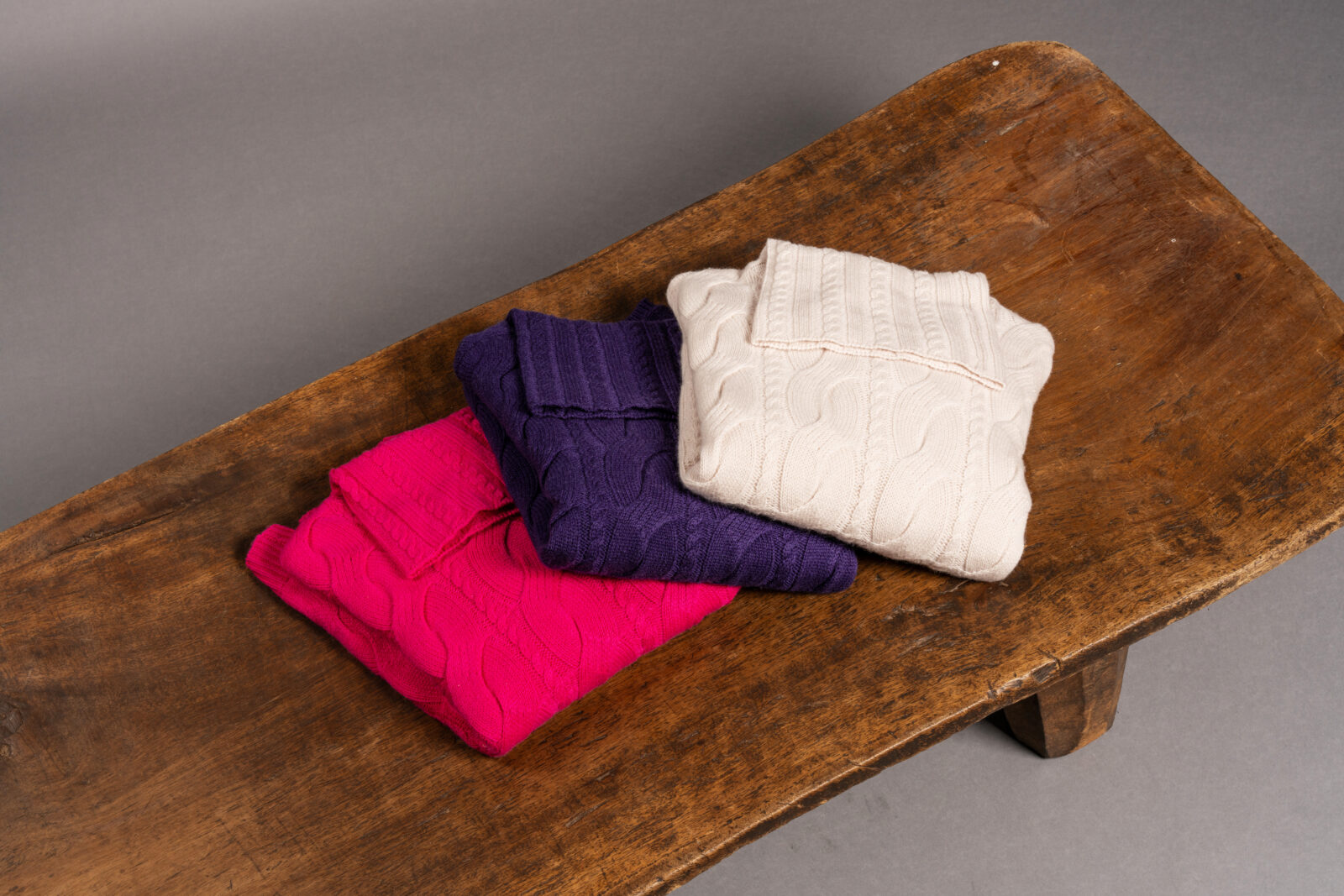 Drei gefaltete Zopfmusterpullover in Creme, Lila und Pink liegen ordentlich gestapelt auf einer rustikalen Holzbank vor einem schlichten grauen Hintergrund.